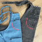 A light blue denim kid's apron folded in half rests on top of an adult's distressed black denim apron, which is also folded in half. Both adults and kids can wear custom-embroidered Cake Hoopla aprons. 