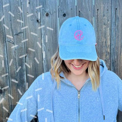 A woman is wearing a light-blue denim hat with a neon pink "CH" embroidered on the front in the center. In the CH logo, the center bar of the H connects to the C, which encircles the H. The logo stands out bright against the blue denim. The denim hat looks good with the woman’s heathered gray zip-up hoodie.  This hat has a ‘90s retro feel contrasted with modern-looking embroidery.