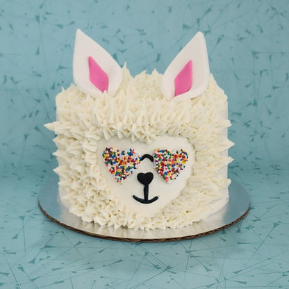 A llama-themed white cake that looks like a llama head with white frosting fur, heart rainbow sunglasses, and pink and white ears; a pre-decorated cake available for shipping or pick up at Cake Hoopla in Tigard, Oregon, in the Portland area.