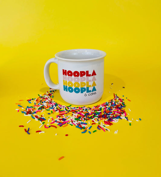The HOOPLA & cake campfire style mug is on a bright yellow background, making the colors pop. Rainbow sprinkles are concentrated around the mug.