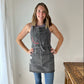 A woman standing 5 feet 9 inches is wearing the black distressed denim apron. The apron covers almost to her neck and extends down to mid-thigh. The straps loop around and are loosely tied in the front around her waist, slightly emphasizing her curves.