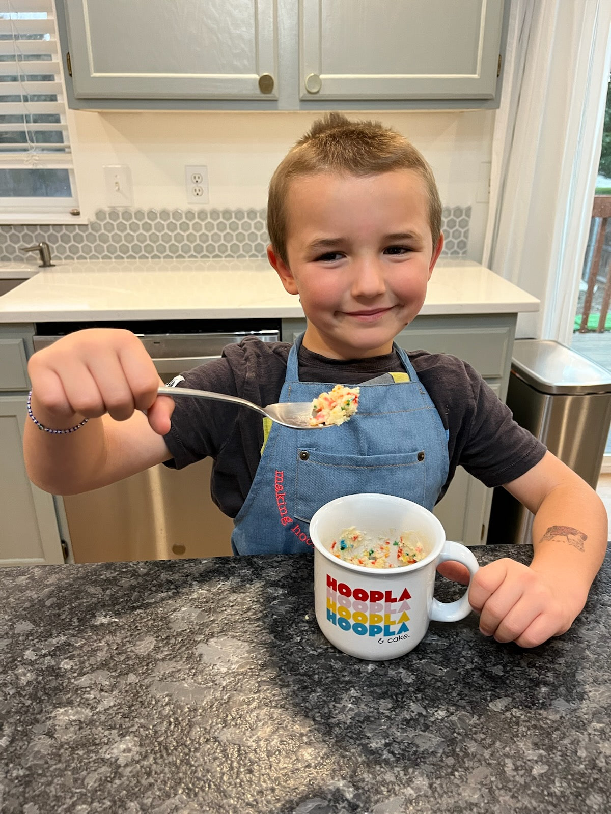 A 5-year-old boy is wearing the making hoopla denim apron as he eats cake from a HOOPLA mug.