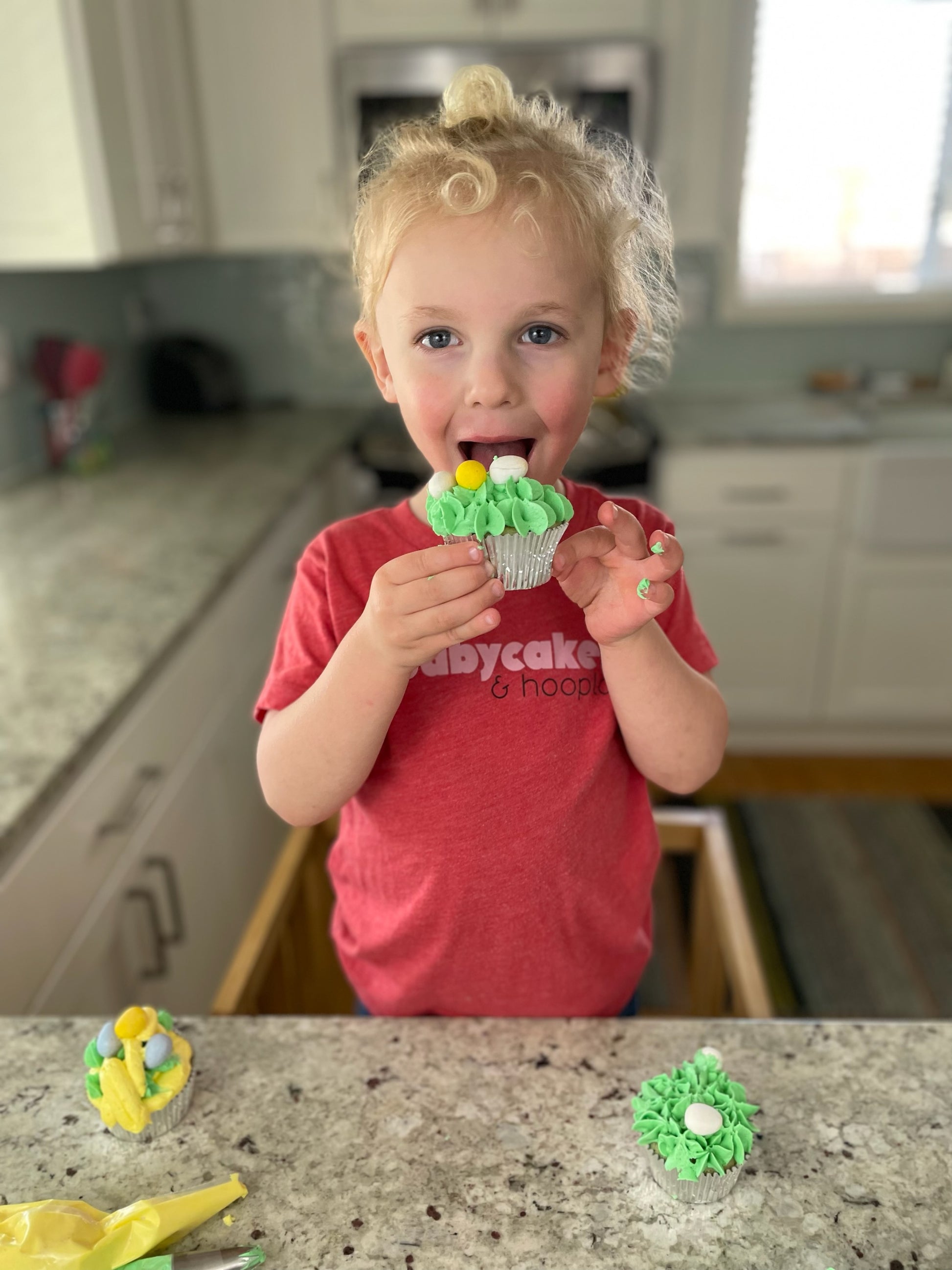 A little boy enjoys the fruits of his labor after taking home a DIY cupcake kit. He smiles as he opens his mouth to eat a cupcake. Two other decorated cupcakes and piping bags lay on the counter in front of him. He is wearing the babycakes and hoopla shirt, which fits him well.