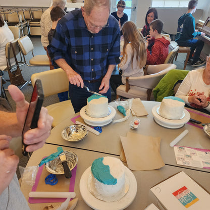 An older man frosts a cake blue during a to-go cake-decorating event at a senior center.