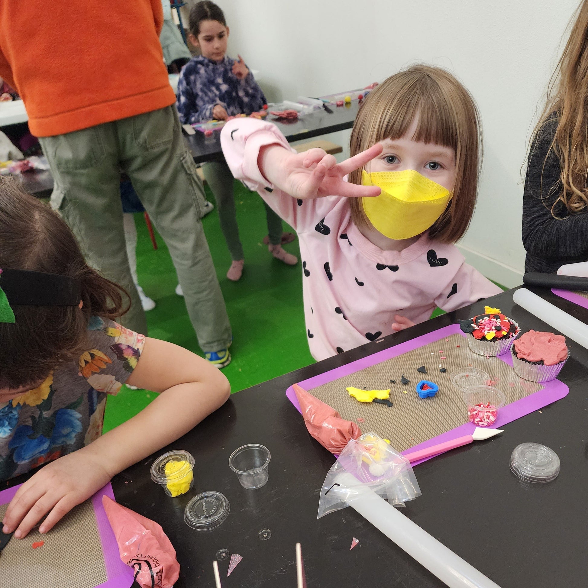A toddler flashes the peace sign at the camera, her partially decorated cupcakes and decorating station on the table in front of her.