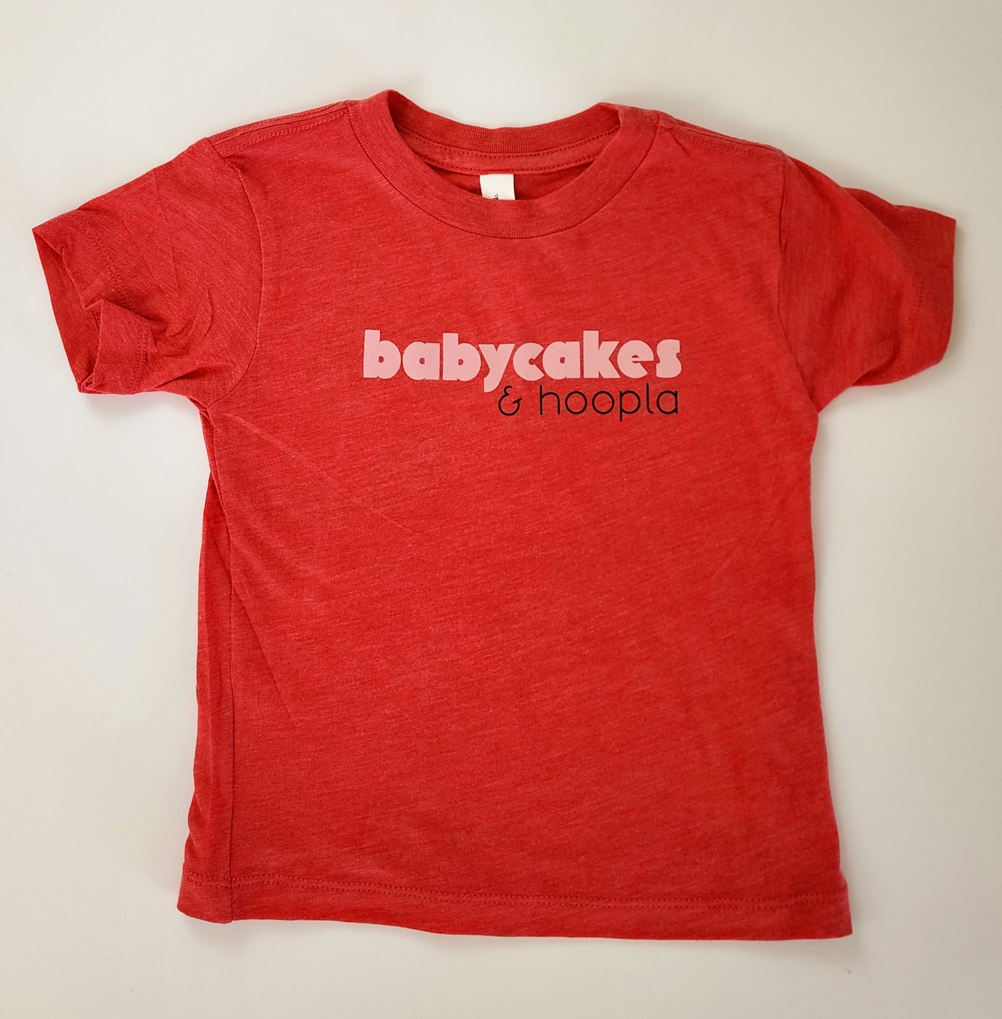 A red t-shirt with a soft, heathered look is spread out on a white background. The front of the shirt reads "babycakes" in a bold, light pink font. Beneath "babycakes" and to the right is "& hoopla" in a thinner black writing.