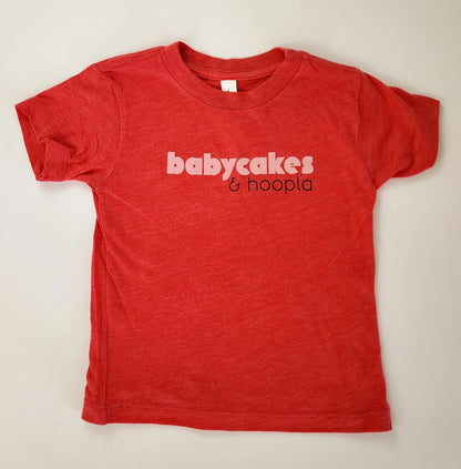 A red t-shirt with a soft, heathered look is spread out on a white background. The front of the shirt reads "babycakes" in a bold, light pink font. Beneath "babycakes" and to the right is "& hoopla" in a thinner black writing.