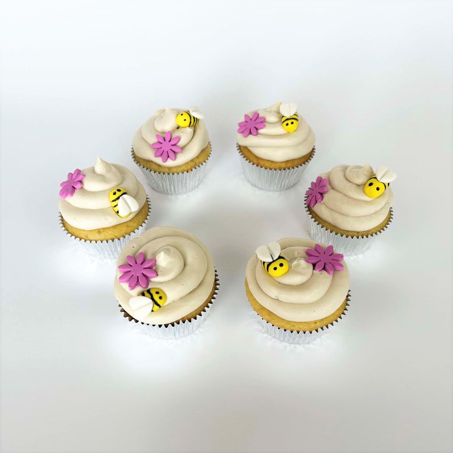 Finished bee-themed DIY cupcakes form a circle, each with a mound of white frosting on top that looks a honeycomb. Pink fondant flowers and yellow, pill-shaped fondant bees with white heart-shaped wings are on top of the frosting. Drawn in black edible marker are two dots for the bees' eyes and three black stripes along the bees' bodies.