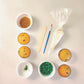 The components of the You’re a Gem cupcake kit, including four of the 12 cupcakes that come with the kit, gold sprinkles, edible green paint dust, a wood spatula, a paintbrush, white frosting in a piping bag, and green rock candy. Also included with this kit but not pictured is an easy-to-follow instruction card. This kit shows the intersection of science and art, making a great STEAM activity.