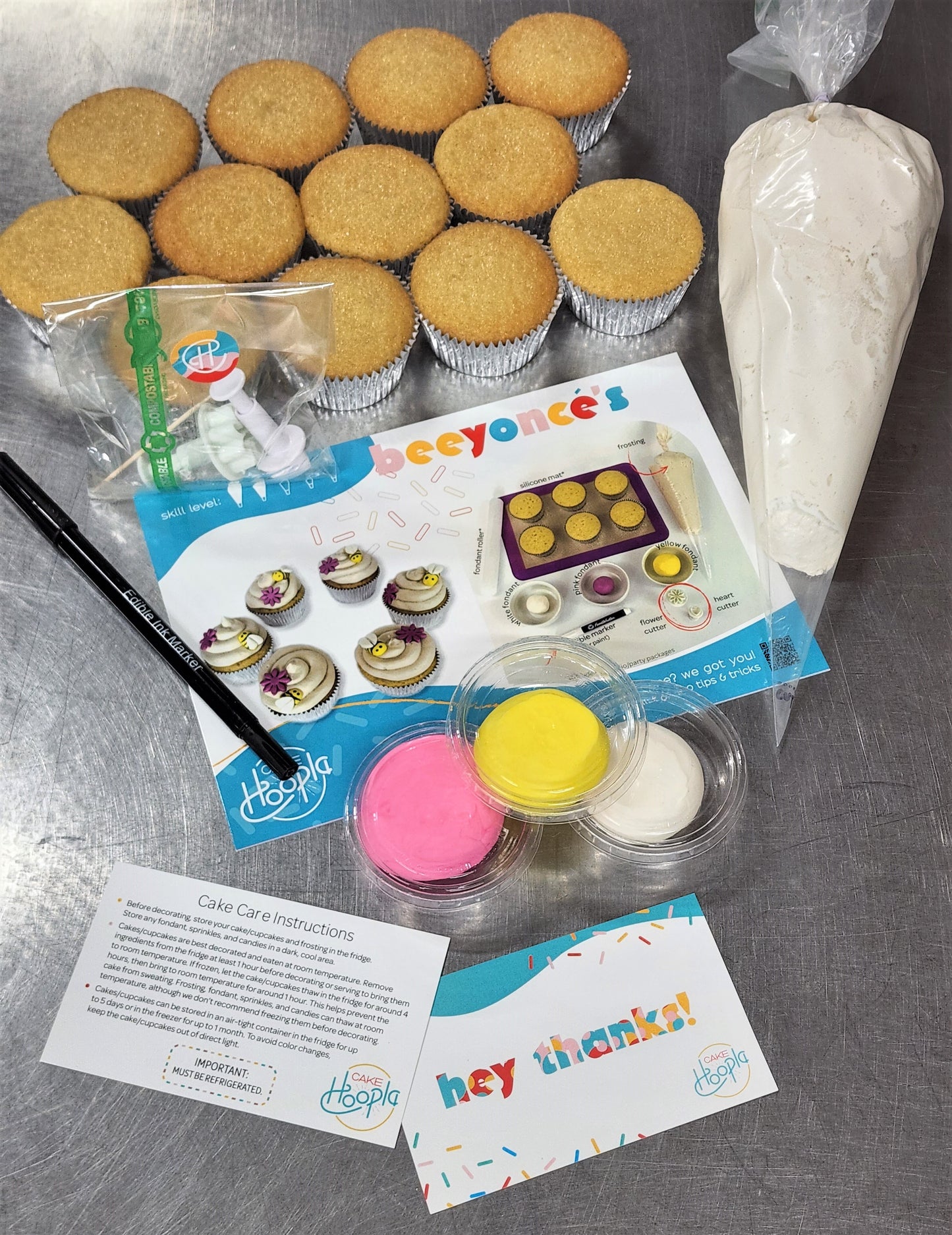 The ingredients and materials in a DIY Beeyonce's cupcake kit. 12 cupcakes, a large piping bag of white frosting, an edible black marker, pink, yellow, and white fondant, two fondant cutters in a compostable cellophane bag, a colorful instruction card with pictures, a cake care instruction card, and a thank you card.