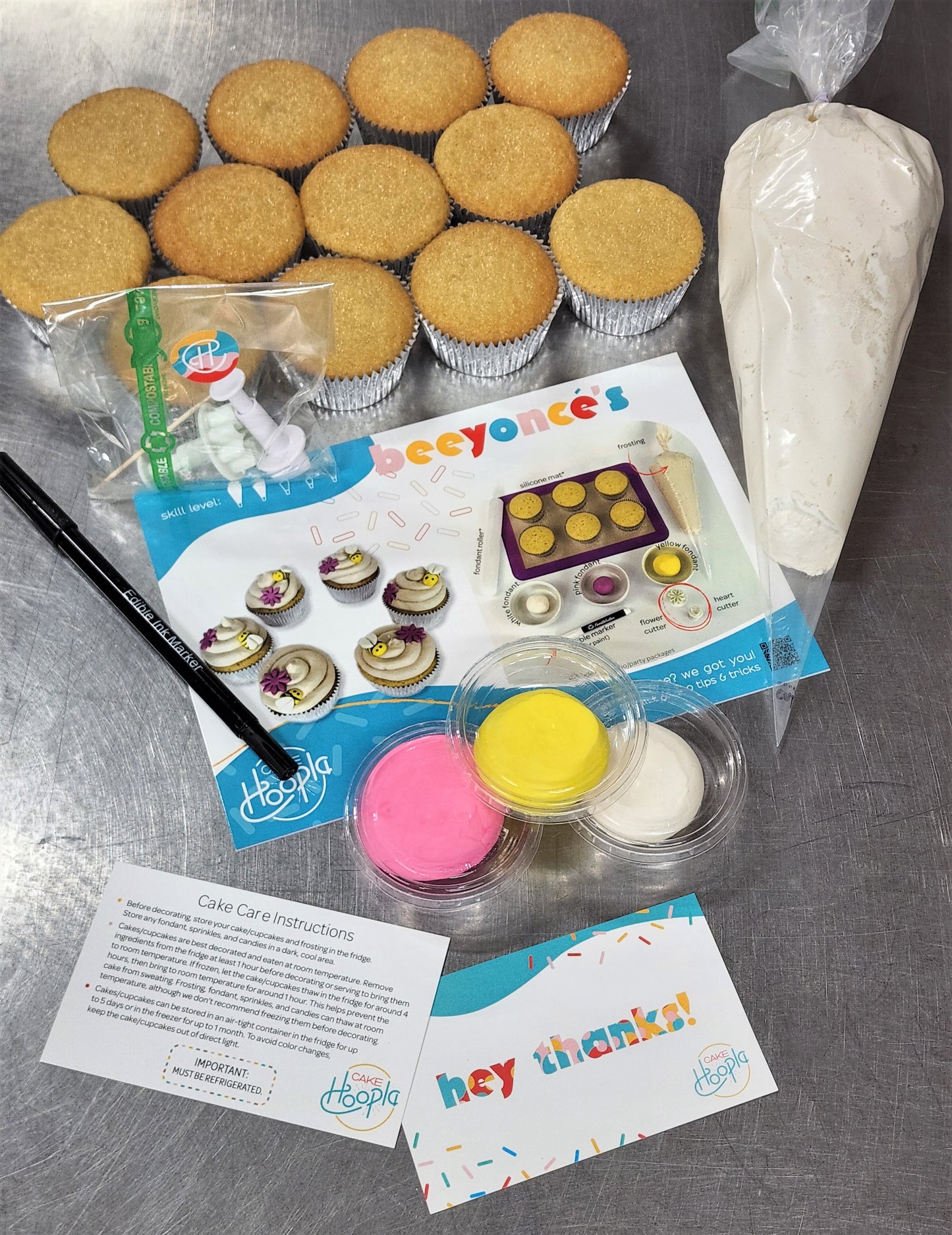 As an example, this shows all the ingredients and materials that go into a cupcake kit. It includes 12 cupcakes, a piping bag with frosting, an edible black marker, pink, yellow, and white fondant, two fondant cutters in a compostable cellophane bag, a colorful instruction card with pictures, a cake care instruction card, and a thank you card. The DIY kits are robust and full of fun components. 