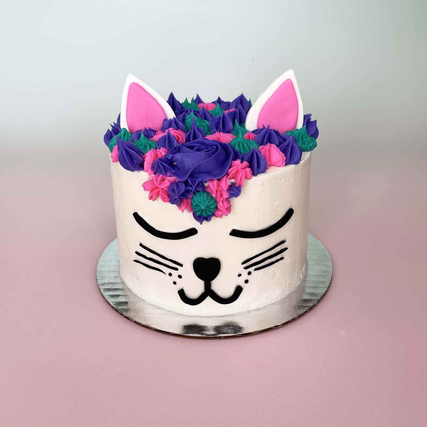 A white cake with a cute cat face decorated in black fondant. The eyes are closed, giving the cat a rested, contented look. The top of the cake is covered in purple, pink, and teal frosting rosettes, stars, and swirls in different shapes and sizes. White fondant ears sit atop the cake and are lined with pink fondant. This fun cat cake is in the genre of unicorns, woodland animals, and other whimsical creatures. 