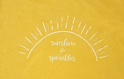 A closeup of the screen printing on the “sunshine & sprinkles t-shirt. “Sunshine & sprinkles” is written in a whimsical, almost cursive font across the center. The words are framed by an abstract half sun with rays of sprinkley sunshine shooting out. 