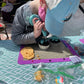 A little girl is at a custom DIY cupcake decorating party in the Portland area. She is concentrating as she pipes blue frosting onto one of two cupcakes. Around her are more decorating tools and ingredients. 