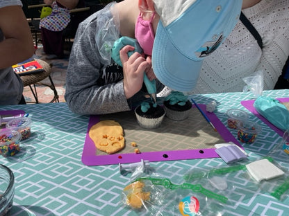 A little girl is at a custom DIY cupcake decorating party in the Portland area. She is concentrating as she pipes blue frosting onto one of two cupcakes. Around her are more decorating tools and ingredients. 