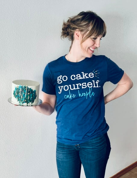 A smiling woman standing 5 feet 9 inches tall and weighing 155 pounds wears the royal blue "go cake yourself” tee. The tee shirt is form fitting, but not too snug, nor too loose. Her upper body is slightly muscular, but the shirt is not too tight against her shoulders or arms. It extends down to the middle of the zipper fly on her jeans, slightly bunching at the hip. 