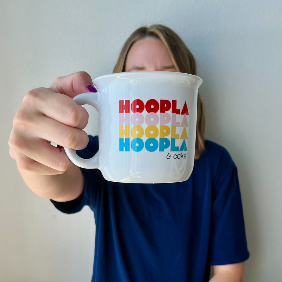 A woman is holding the handle of a large white, shiny, campfire-style ceramic mug. It has the word "HOOPLA" written 4 times, one word in each row, and in all caps. The font has a slightly retro feel. Each HOOPLA has a different color: red, pink, yellow, and teal. Under the 4 HOOPLAs and to the right are "& cake" in a simple black font. This 15oz mug is larger than a standard mug and has bright, contrasting colors. 