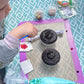 A toddler is at a custom DIY cupcake party in the Portland area and has two cupcakes in front of her on top of a silicone mat, along with sprinkles and a piping bag filled with blue frosting. She is concentrating on painting the cupcakes.