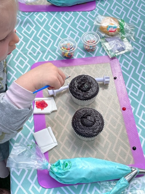 A toddler is at a custom DIY cupcake party in the Portland area and has two cupcakes in front of her on top of a silicone mat, along with sprinkles and a piping bag filled with blue frosting. She is concentrating on painting the cupcakes.